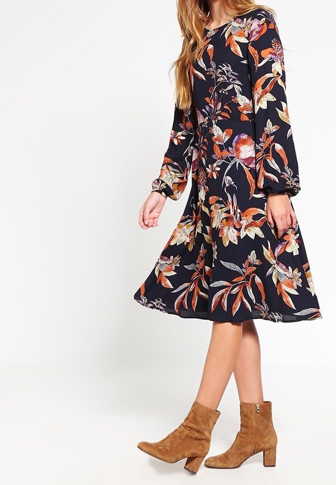 Robe imprimée florale avec des manches longues, corsage ajusté et jupe évasée ; fond marine avec des fleurs orange, rose et vert ; assortie à des bottines beiges.