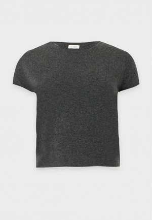 Une femme porte une chemise à manches courtes gris charbon, un pantalon marron à jambes larges avec une ceinture et des chaussures plates foncées, sur un fond uni.
