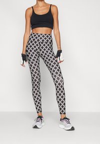 Černý crop top s tenkými ramínky, doplněný o legíny s vysokým pasem s geometrickým černobílým vzorem. Bílé sportovní boty s růžovými detaily.