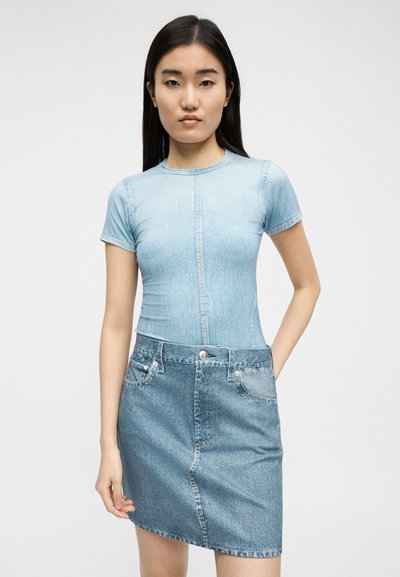 Νεαρή γυναίκα με μακριά σκουρόχρωμα μαλλιά, ντυμένη με ένα στενά εφαρμοστό τζιν μπλουζάκι σε ανοιχτό μπλε χρώμα και μια ψηλόμεση τζιν φούστα με τσέπες.