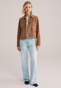 Bruine suède cropped jas met twee voorzakken, knoopsluiting en een kraag, gecombineerd met lichtblauwe flare jeans en zwarte schoenen.