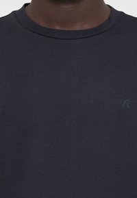 Μπλε βαμβακερό T-shirt με κανονική λαιμόκοψη, διαθέτοντας μια διακριτική ανάγλυφη λογότυπο σε ταιριαστό χρώμα στην αριστερή πλευρά του στήθους. Λείος υφασμάτινος χαρακτήρας.