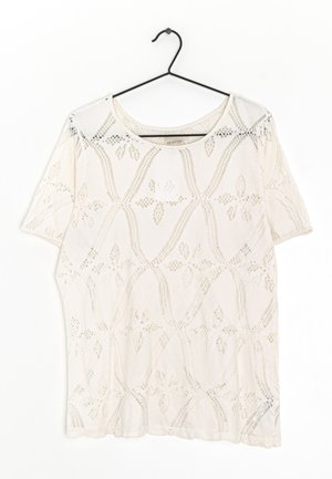 Haut blanc à manches courtes avec col rond et motif en dentelle florale géométrique, présenté sur un cintre noir contre un fond blanc.