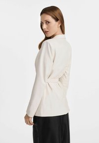 Biała dopasowana marynarka z długimi rękawami, z detalem w postaci paska w talii oraz gładkim, strukturalnym wykończeniem, noszona na czarnej odzieży.