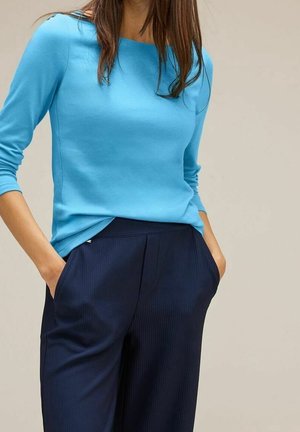 Femme portant un haut à manches longues bleu vif rentré dans un pantalon ample plissé bleu marine foncé, les mains dans les poches, sur un fond neutre.