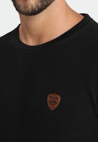 Zbliżenie na dolną część twarzy i szyję mężczyzny ubranego w czarną teksturowaną koszulę z małym brązowym logo w kształcie tarczy na piersi.