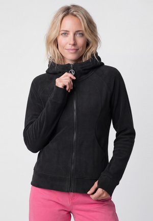 Blonde Frau, die einen schwarzen Hoodie zumacht, trägt pinke Hose und steht vor einem einfarbigen hellen Hintergrund.