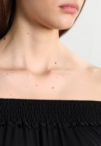 Haut noir à épaules dénudées avec encolure smockée, tissu léger et bordure à volants. Peau avec des grains de beauté visibles sur le cou en dessous.