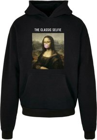 Fekete kapucnis felső, elülső grafikával, amelyen a Mona Lisa látható egy rózsaszín buborékfóliával, felette a szöveg: "A KLASSZIKUS SZELFIE."
