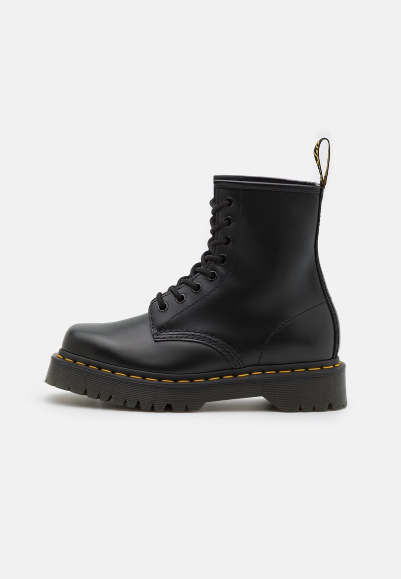 Botte de combat noire en cuir à lacets avec semelle épaisse en caoutchouc, coutures jaunes et languette à la talon sur un fond uni clair.