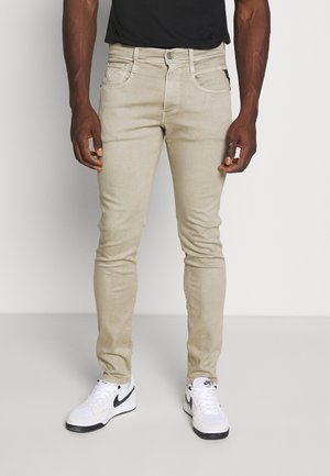 Mand iført slanke beige bukser og hvide Nike sneakers står mod en ensfarvet lys baggrund.