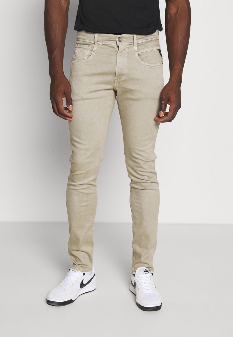 Homme portant un pantalon beige slim et des baskets blanches Nike, debout devant un fond clair uni.