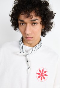 Hvit bomulls T-skjorte med et rødt solmotiv på brystet. Et svart-hvitt bandana med prikker er knyttet rundt halsen.