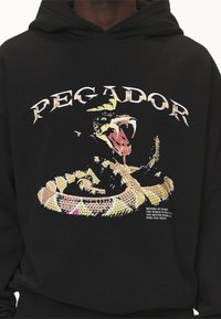 Černá mikina s kapucí s grafikou žlutého hada s detailní hlavou a zuby, texturovaným designem a bílým nápisem nahoře a dole.