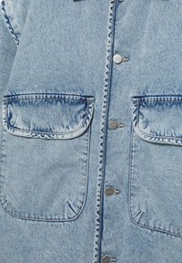 Hellblaues Denim-Oberteil mit zwei großen Klappentaschen und vier silbernen Knöpfen, die vertikal in der Mitte verlaufen.