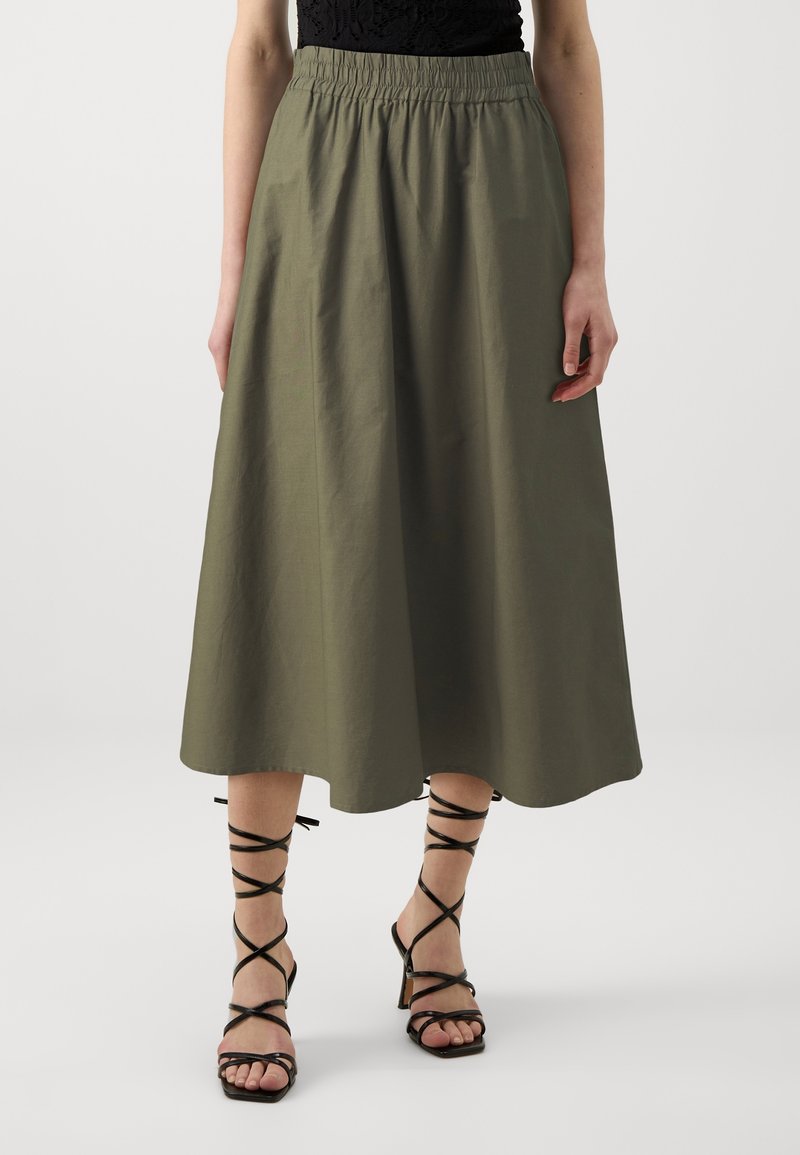 Femme portant une jupe midi vert olive à taille froncée et des sandales à talons hauts noires à lacets, debout devant un fond uni.