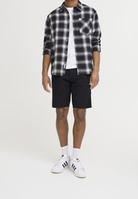 Chemise en flanelle à carreaux noir et blanc, t-shirt blanc, shorts noirs et baskets blanches avec des rayures noires. Comprend des boutons en laiton et une poche poitrine.