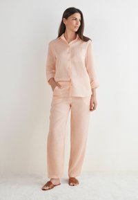 Ljust rosa linneskjorta med långa ärmar och framsida av knappar, ihop med matchande löst sittande byxor. Bruna sandaler fulländar outfiten.