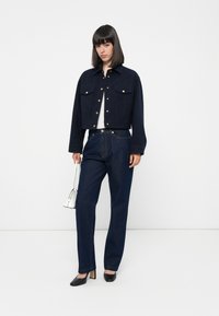 Navy cropped jacket with two front pockets and gold buttons, paired with dark denim trousers. Model holds a small white bag and wears black heels.
