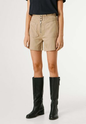 Mujer con pantalones cortos beige de tiro alto con cremallera y botones frontales, combinados con botas de cuero negras hasta la rodilla y una blusa negra.