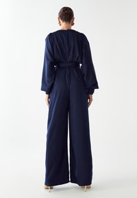 Marineblauwe jumpsuit van een gladde stof met lange mouwen, een ceintuur in de taille en wijde broekspijpen. Achterste ritsluiting.