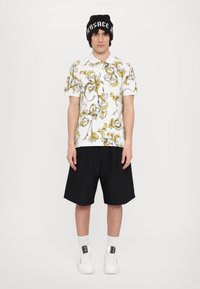 Biała koszulka polo z żółtymi i czarnymi ozdobnymi wzorami, zestawiona z czarnymi szortami. Model ma na sobie czarną czapkę beanie i białe sneakersy.