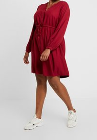Vestito rosso a maniche lunghe con scollo a V e rifiniture in pizzo, stretto in vita da un laccetto. Indossato con sneakers bianche.