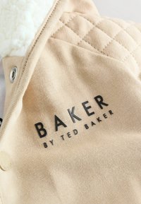Bež vooderdatud riie, millel on kontrastne valge, karvane vooder. Omab ümmargust nööpainet ja trükitud brändingut: "BAKER BY TED BAKER."