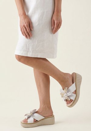 Frau trägt weiße Keilsandalen mit Blumenverzierung und ein weißes knielanges Kleid, steht mit einem leicht angehobenen Bein.