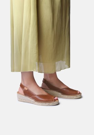 Woman wearing light brown leather wedge sandals with open toes and ankle straps, paired with a flowing pale yellow skirt.