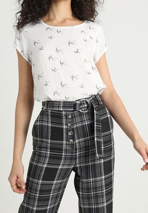 Vrouw draagt een wit kort mouwtopje met vogelprints en een hoog getailleerde zwart-grijze geruite broek met knopen en een stoffen riem.