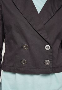 Gros plan d'un blazer noir croisé avec quatre boutons argentés, porté sur un top bleu clair.