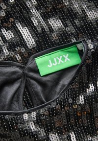 Mustekki musta paljettikangas kiiltävällä pinnalla, jossa on vihreä etiketti, jossa lukee "JJXX". Sisäosa on sileä ja musta, ja siinä on tiukka muotoilu.