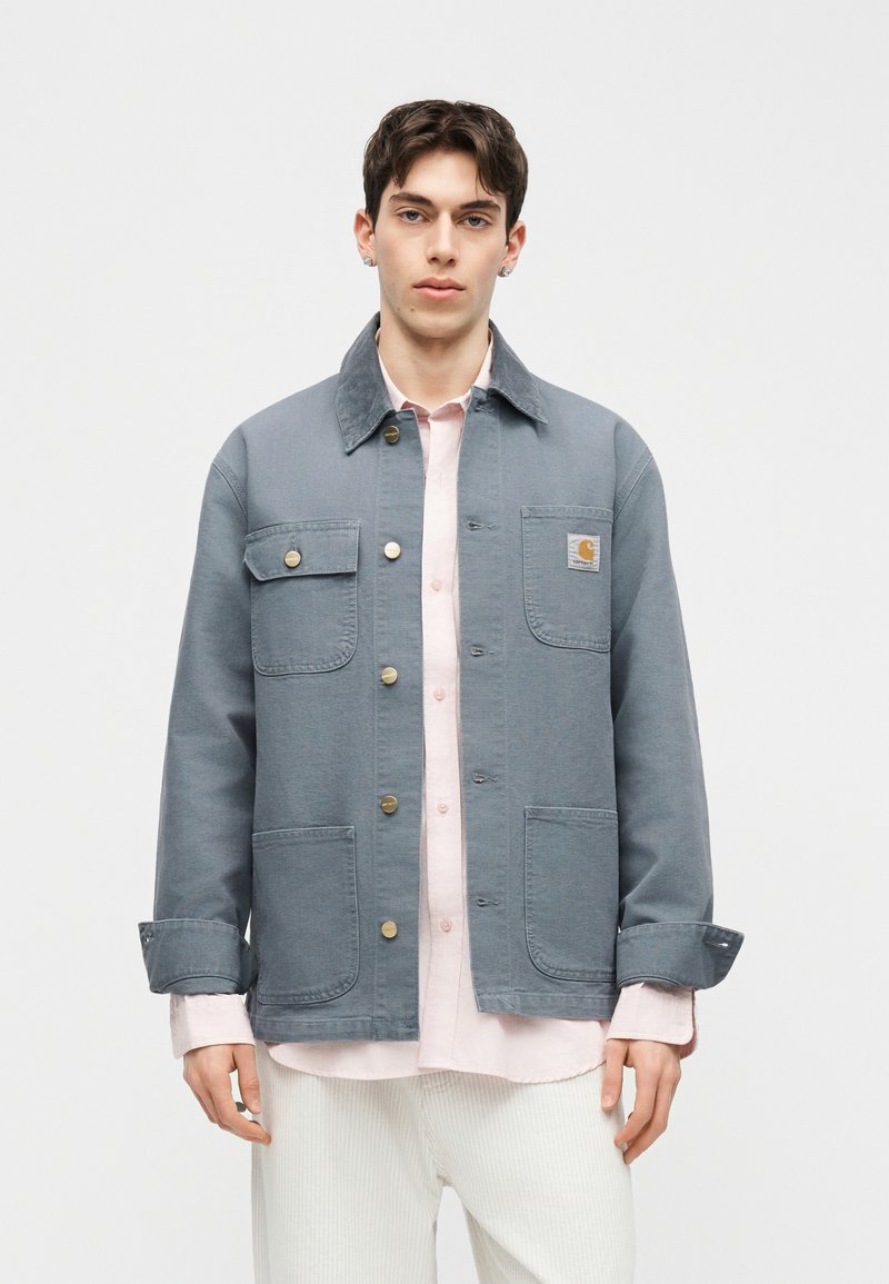 Giovane uomo con giacca blu tenue abbottonata sopra una camicia rosa chiaro e pantaloni bianchi, in piedi davanti a uno sfondo semplice.