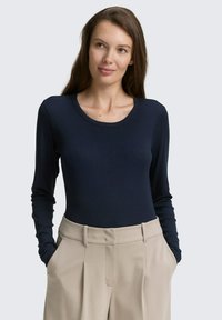 Kvinna med lång brunt hår bär en marinblå långärmad skjorta instoppad i beige högmidjade byxor med händerna i fickorna, ser något åt höger.