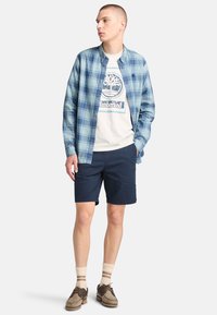Camicia a maniche lunghe a quadri blu, t-shirt grafica bianca, pantaloni corti blu scuro, calze crema con strisce marroni e scarpe marroni, in posa in piedi.