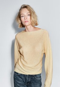 Frau mit kurzen blonden Haaren, die einen beigen Pullover und blaue Jeans trägt, steht vor einem schlichten hellgrauen Hintergrund.