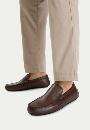 Piernas con pantalones beige que llevan mocasines marrones de cuero con detalles de costura y suelas de goma sobre un fondo blanco.