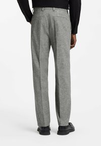 Homme portant un pantalon gris coupe droite, une chemise noire boutonnée et des mocassins noirs, vu de dos sur fond blanc.