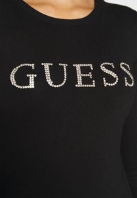 Pull noir à col rond avec le mot « GUESS » écrit en larges strass brillants sur la poitrine.