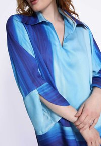 Blouse à dégradé bleu avec col, présentant un design fluide et des manches larges. Le tissu a une texture lisse et des variations de couleur subtiles.