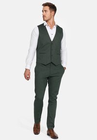 Mørkegrønn treskjøtet dress laget av teksturert stoff, med vest med knapper, kombinert med en hvit skjorte og brune pen-sko.