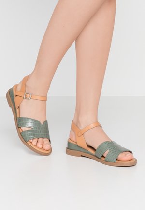 Voeten met groene, gestructureerde en beige leren sandalen met open tenen en enkelbandjes op een lichtgrijze vloer en achtergrond.
