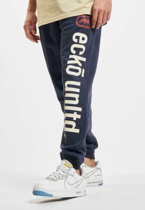 Homme portant un jogging bleu marine avec le logo « ecko unltd » en grandes lettres blanches sur la jambe gauche, associé à des baskets blanches avec des semelles bleues.