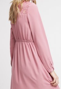 Langærmet pink kjole med blonder på yoken, elastisk talje og sammensyning. Blødt stof med et svævende design og knaplukning ved ærmerne.