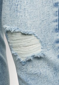 Los jeans de mezclilla azul claro presentan un gran parche rasgado en la rodilla con bordes deshilachados, que revela un forro blanco debajo.