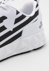 Bílé tenisky s černým pruhovaným vzorem; texturovaný svršek, měkké tkaničky a sochařská, geometrická bílá podešev s černými akcenty.