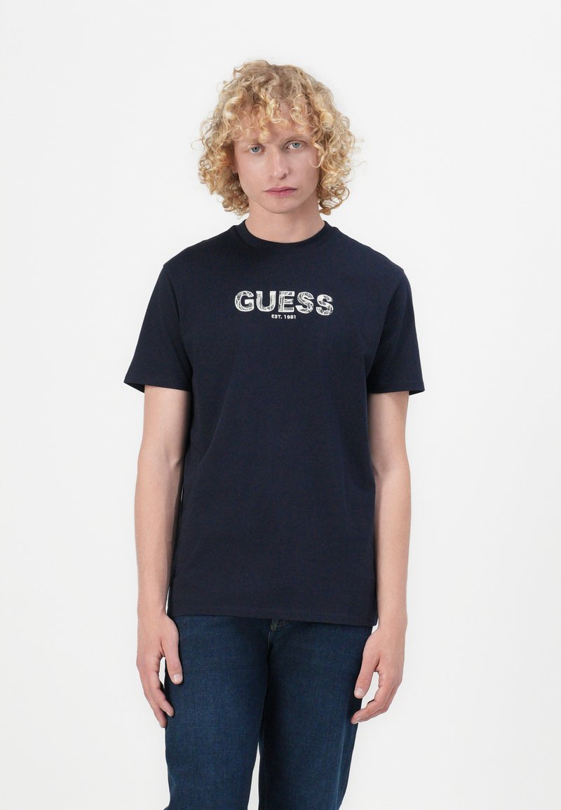 Námornícky modré bavlnené tričko s krátkymi rukávmi, so stredovým bielym grafickým logom "GUESS EST. 1981" na hrudi. Hladká textura.