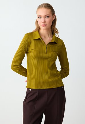 Kobieta w musztardowej prążkowanej bluzce z długim rękawem, z zamkiem przy kołnierzu, w ciemnobrązowych spodniach, stojąca na jednolitym tle.