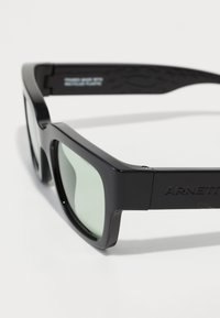 Mustad must plastikprillid, millel on ristkülikukujulised läätsed, rohelise tooniga läätsed ja tekstuuriga mustad templid, millel on "ARNETTE" logo.